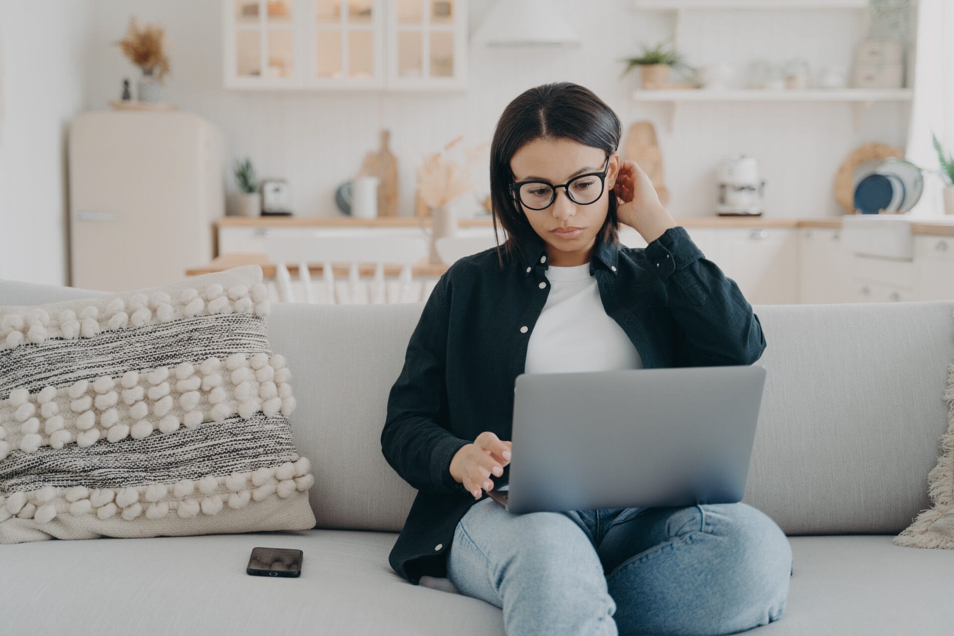 Young woman with glasses sitting with her laptop on sofa at home Woman with glasses using a laptop for online therapy while sitting cross-legged.