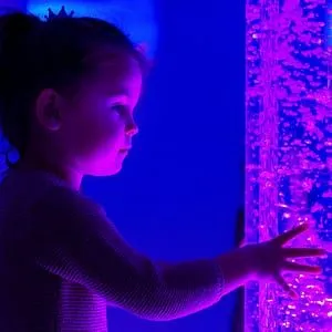 A young girl in a sensory room reaching out to touch a free-standing bubble tube - representing the Purple House Clinic's sensory assessment service.