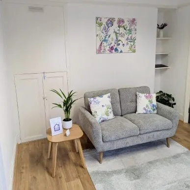 Beautiful white-walled therapy room with Victorian features, a contemporary grey woollen sofa, small table and green plant. A safe and comfortable setting for psychological therapy sessions at the Purple House Clinic Nottingham.